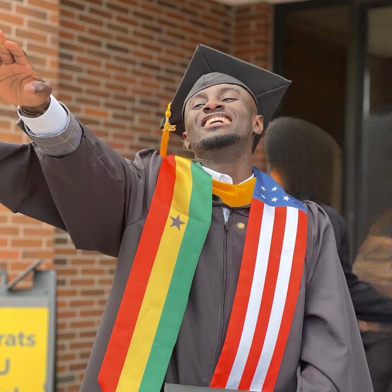 A Ghanaian graduate in cap and gown wearing Ghana and United States stoles, holding a diploma